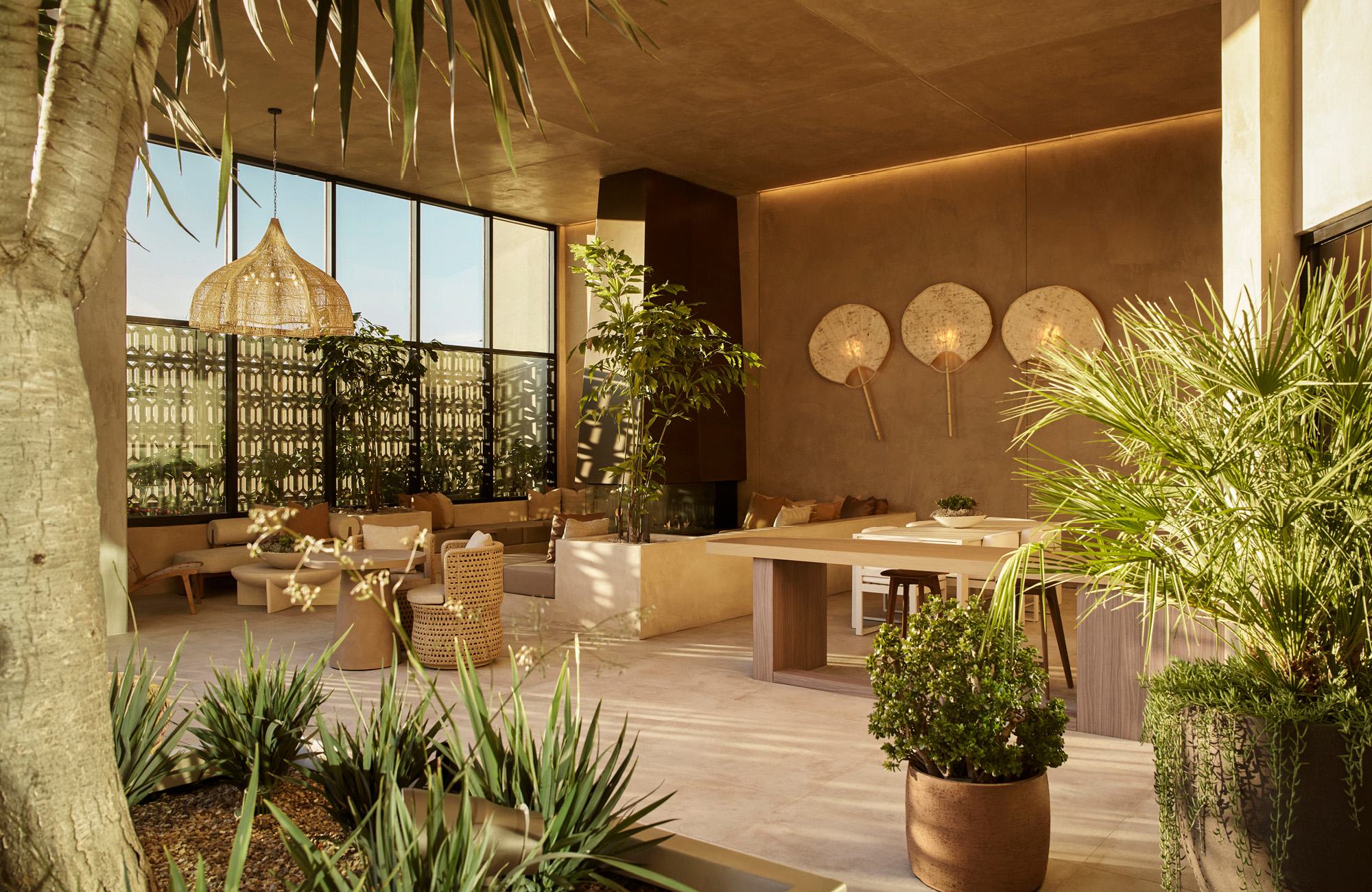 A sunlit, modern outdoor lounge at Rowyn featuring warm earth tones, cozy seating, huge floor-to-ceiling windows, a fireplace, and abundant potted tropical plants.