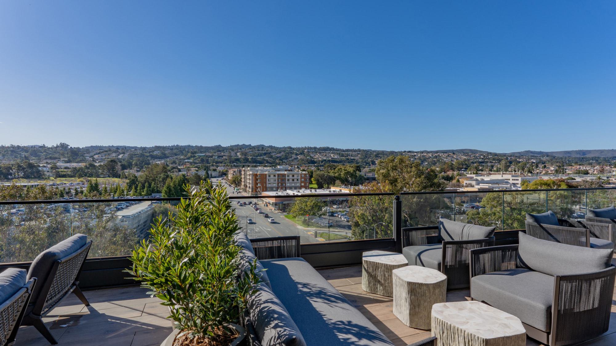 An upscale outdoor rooftop patio with modern lounge seating overlooking a tree-lined street and the hills of Burlingame under a clear blue sky.