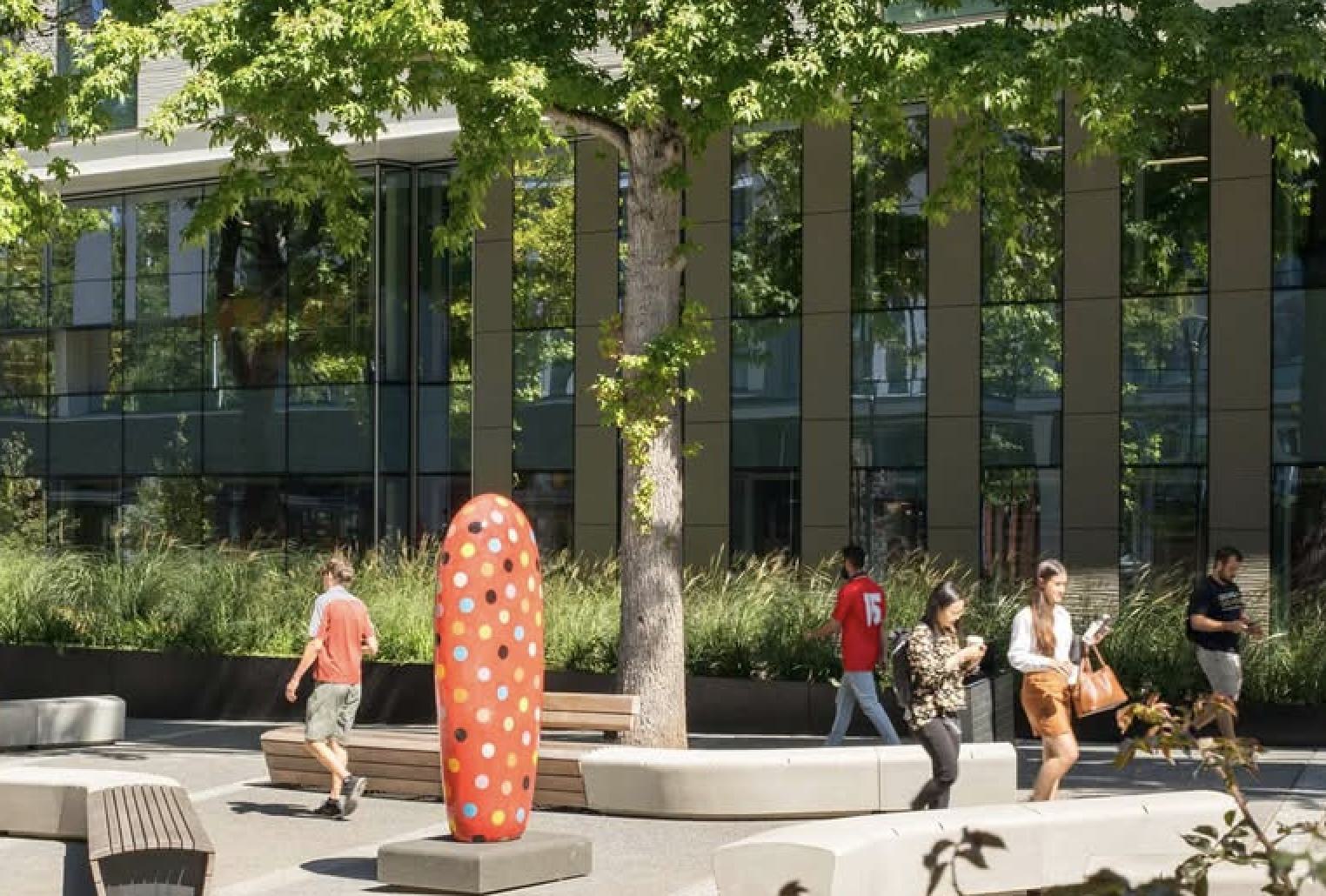 Pedestrians walking through a sunlit plaza in South Lake Union, featuring green trees, a large polka-dotted modern art sculpture, and a contemporary glass-windowed corporate building in the background.