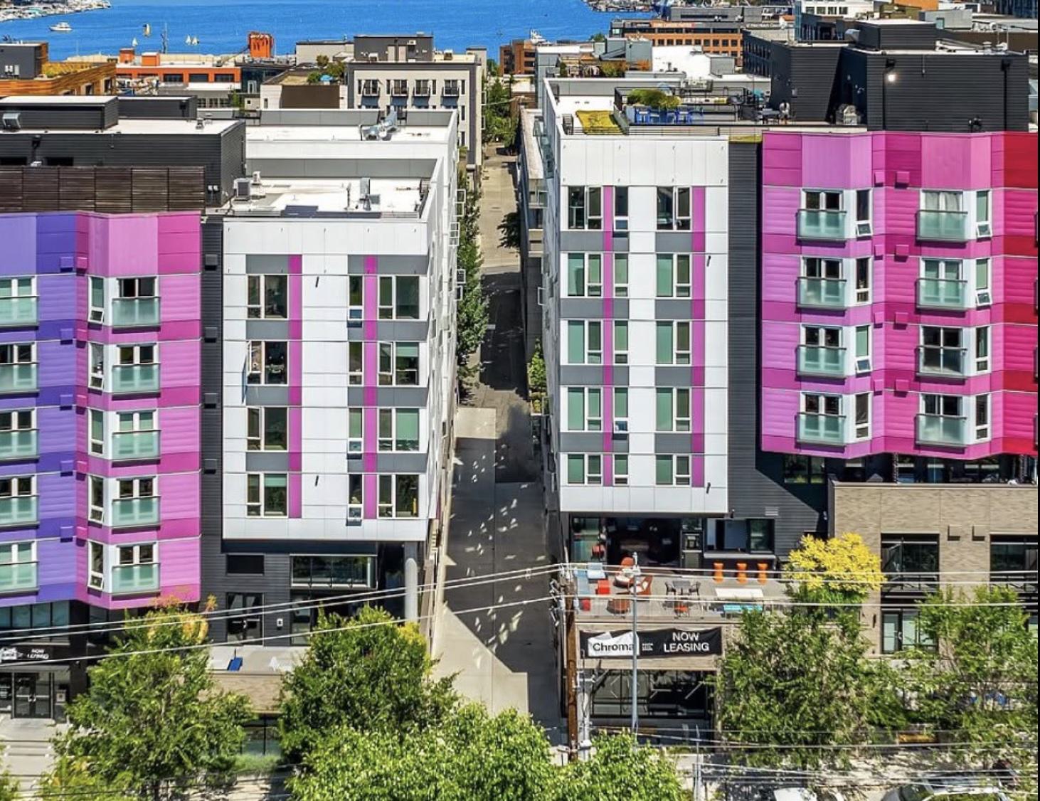 High-angle exterior view of Chroma Apartments in Seattle, showcasing a modern multi-story residential building with vibrant pink, purple, and white facade accents, an outdoor patio plaza, and Lake Union visible in the background.