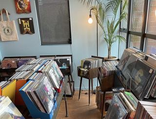 An interior view of the record store section inside Empire Roasters & Records, showing wooden bins, tables, and crates packed with vinyl records next to a large window, a floor lamp, and a potted palm tree.