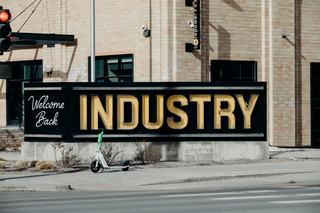 Large black sign reading "Welcome Back INDUSTRY" with a lime green electric scooter parked in front of a brick building in Denver's RiNo district.