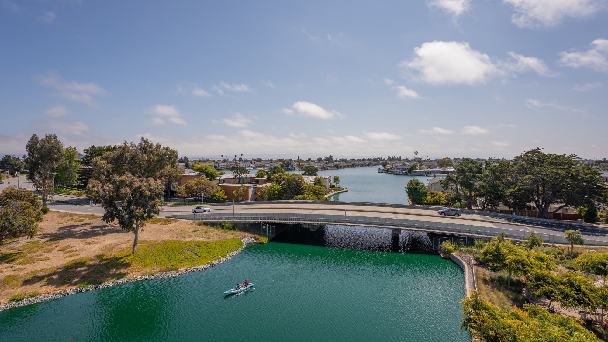 Aerial view of a kayaker paddling through the Foster City lagoon beneath a road bridge.