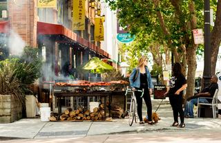 Jack London Square street scene with sidewalk barbecue, cyclists, and tree-lined storefronts.