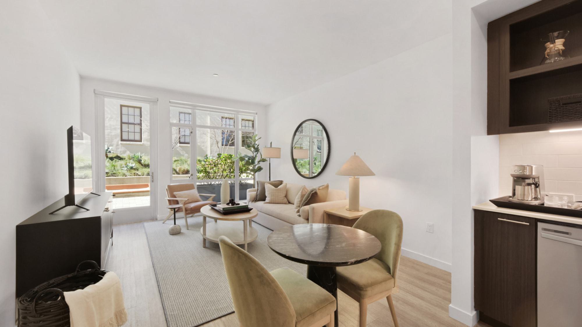 Modern open-concept living area at Alchemy Apartments featuring neutral-toned furniture, a small dining table, wall-mounted TV, and large, oversized glass patio doors looking out to a landscaped terrace.