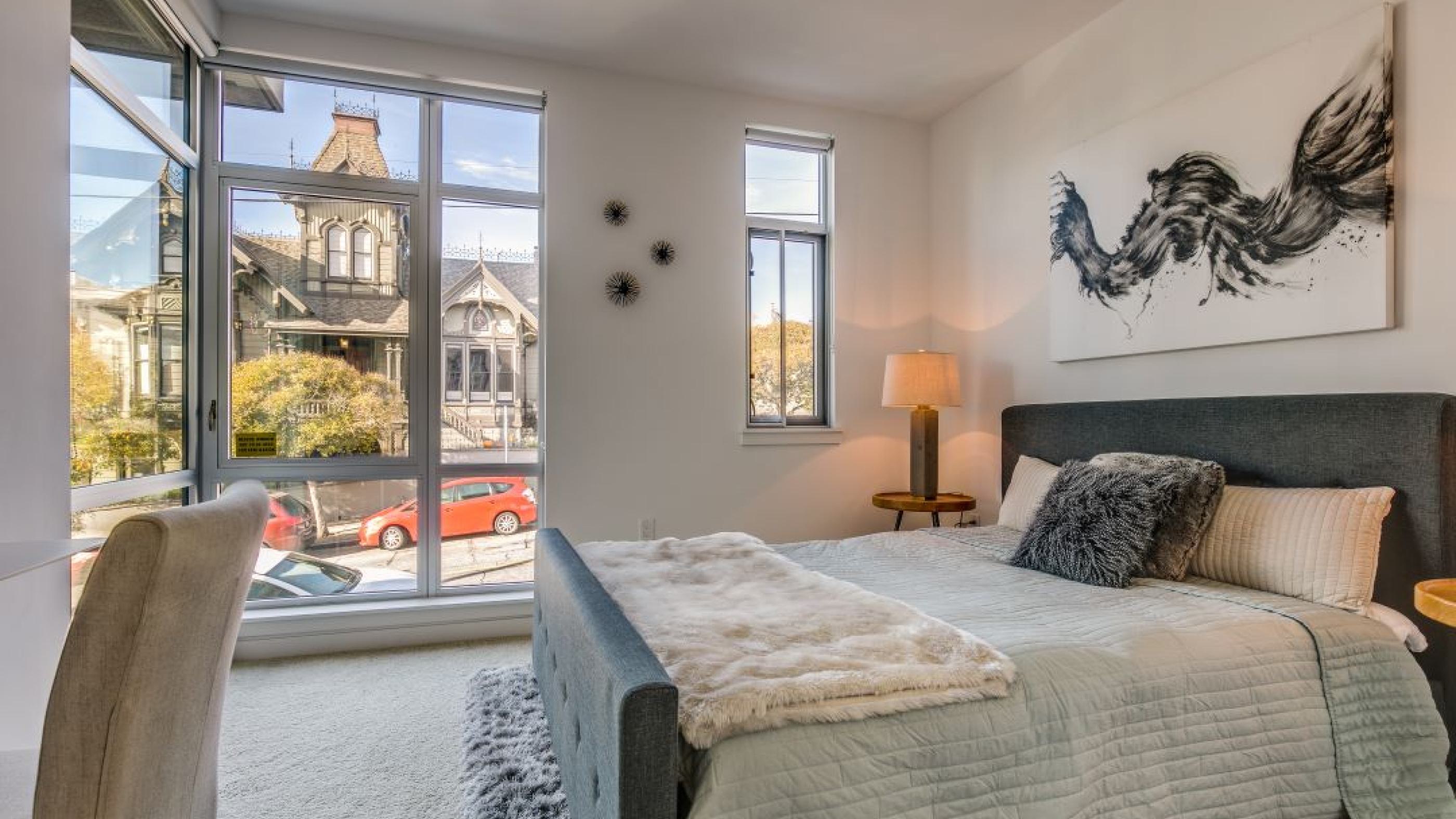 Sunlit apartment bedroom at Alchemy with a dark padded bed frame, abstract wall artwork, and large floor-to-ceiling windows overlooking Victorian homes in Hayes Valley.