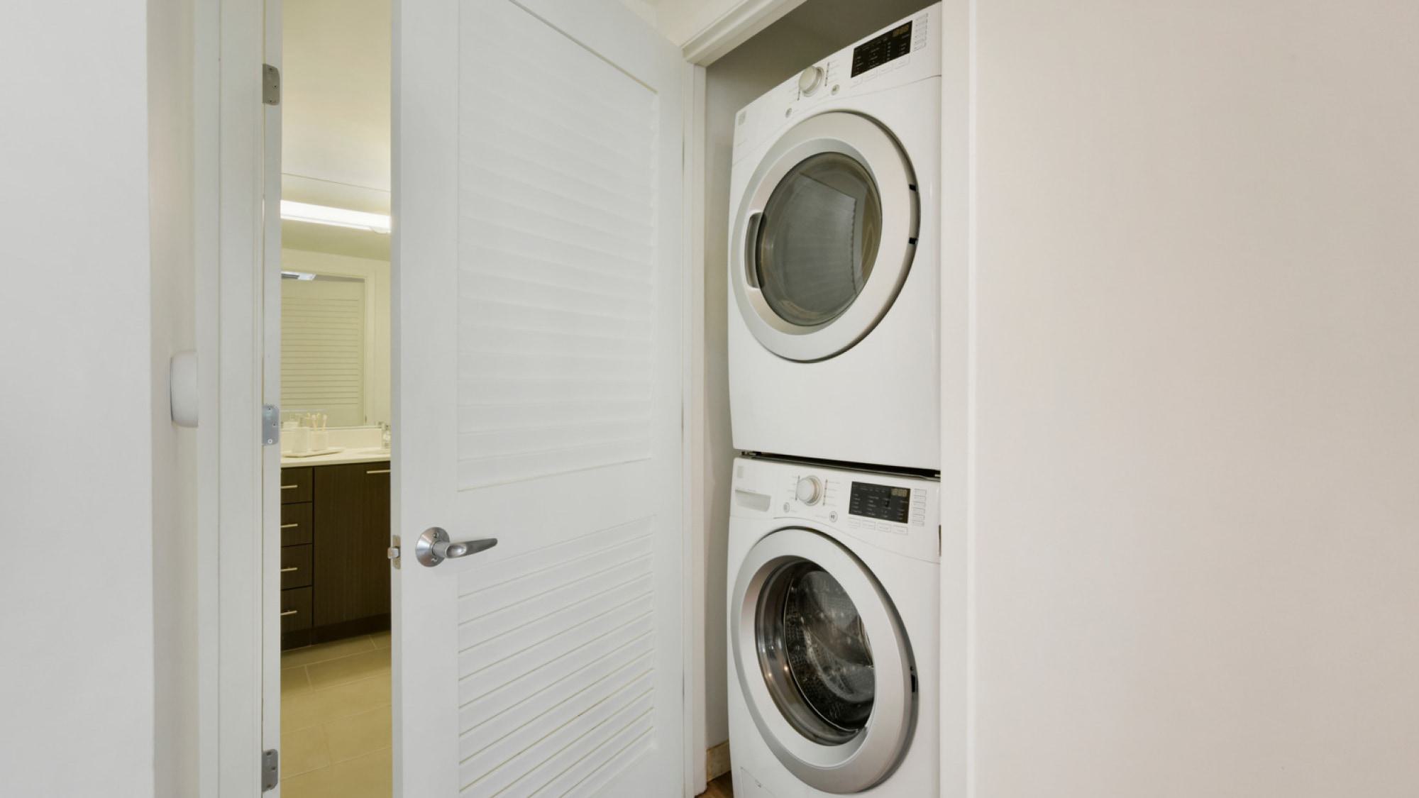 A white, stacked front-loading washer and dryer inside an in-unit closet at Alchemy Apartments, located next to an open bathroom door.