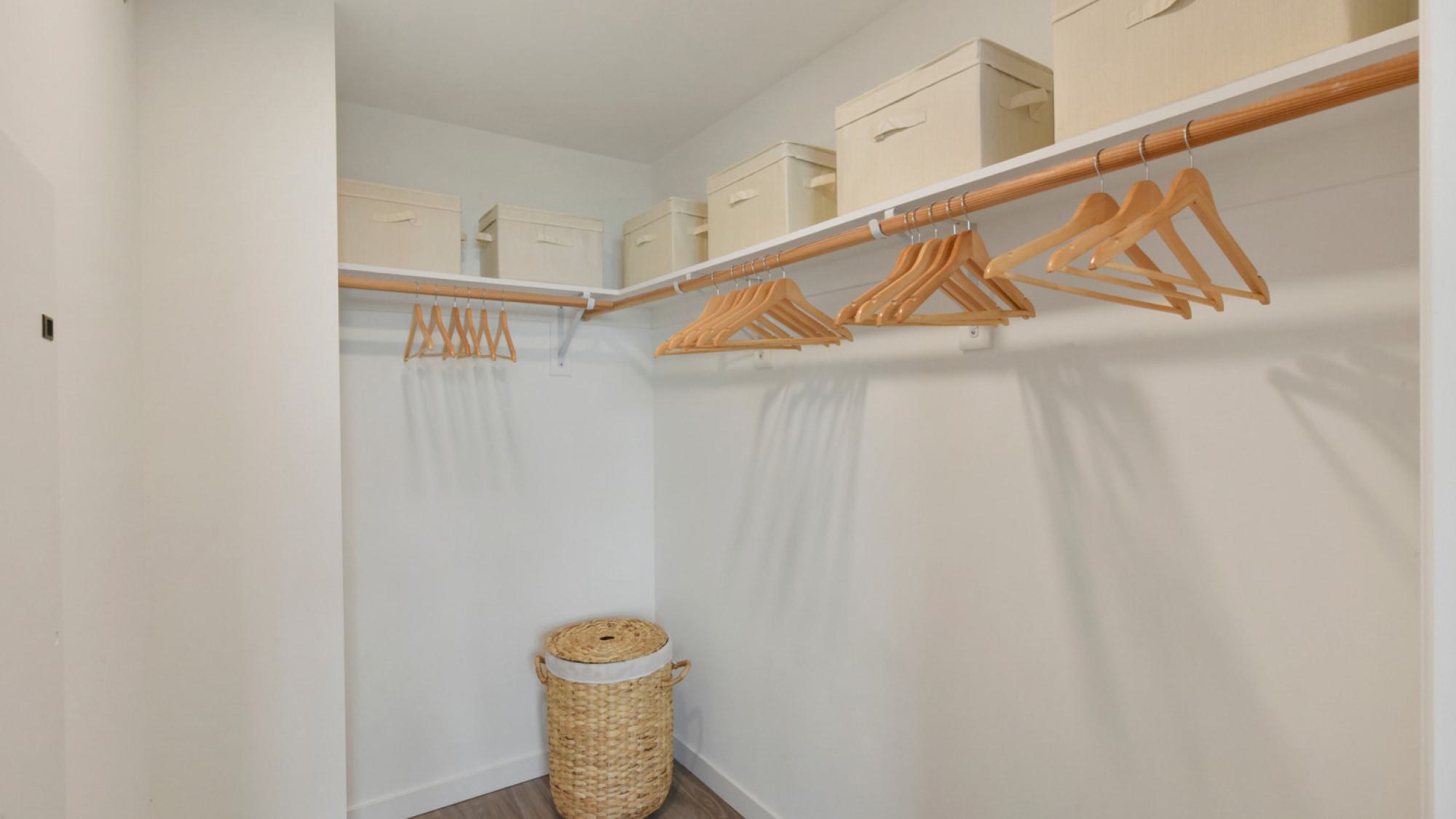 A spacious walk-in closet in an apartment at Alchemy, featuring built-in wooden hanging rods, top shelving with storage boxes, and a woven laundry hamper.