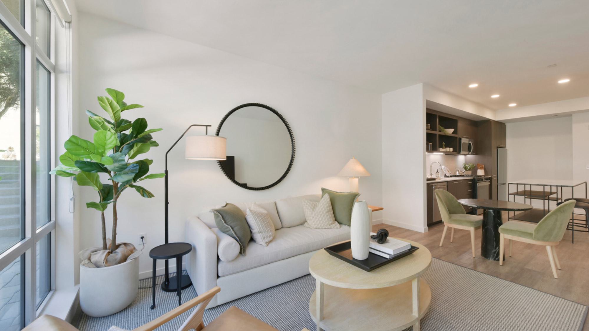 Modern apartment living room at Alchemy featuring a light sofa, large floor-to-ceiling windows, and an adjacent dining and kitchen space.