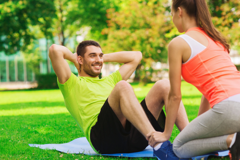 a guy in green shirt & black sport shorts doing sit ups while a girl with pony tail holds his feet in a park - lax apartments