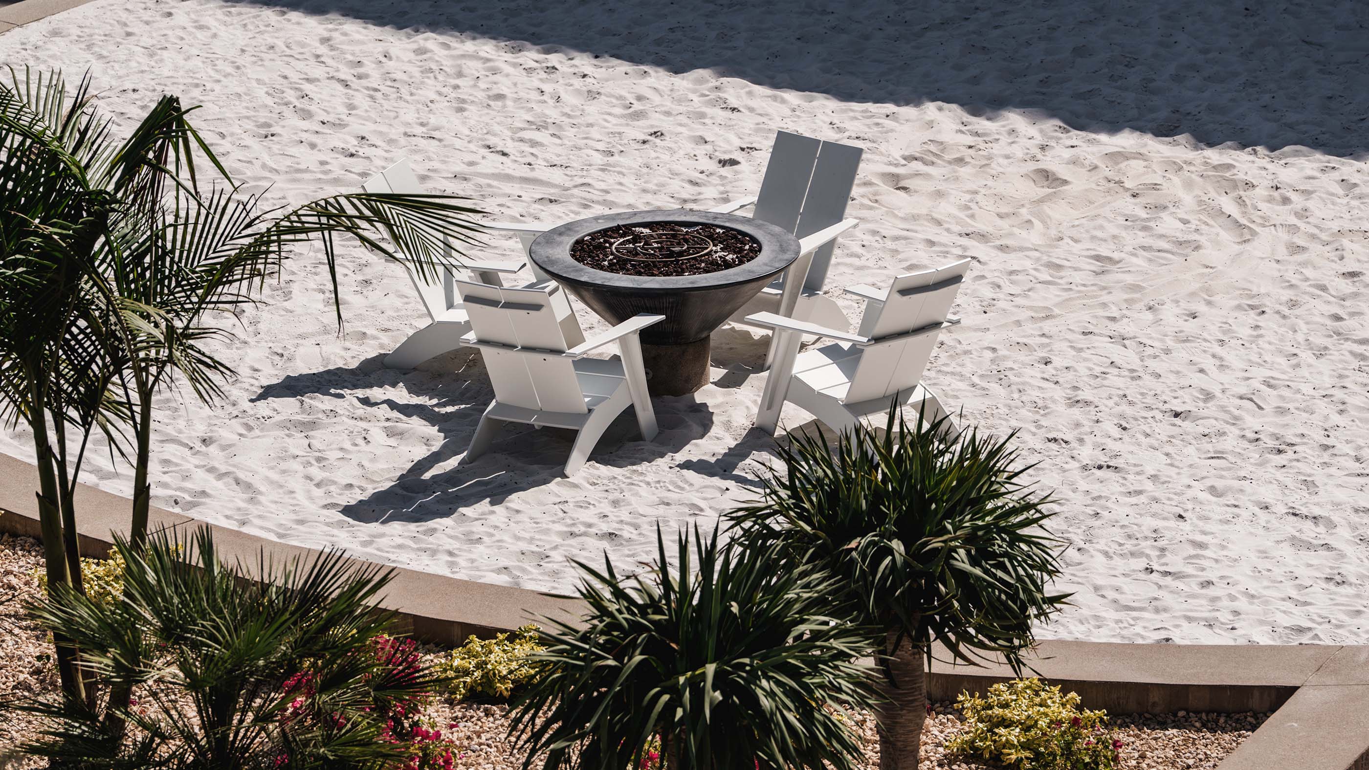 Sandy private courtyard at Stella Apartments featuring white Adirondack chairs around a fire pit surrounded by palm trees.