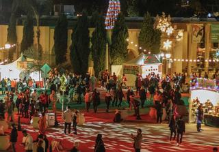 Crowds gathering at Balboa Park December Nights surrounded by festive light displays, food vendor tents, and a tall illuminated Christmas tree in the background.