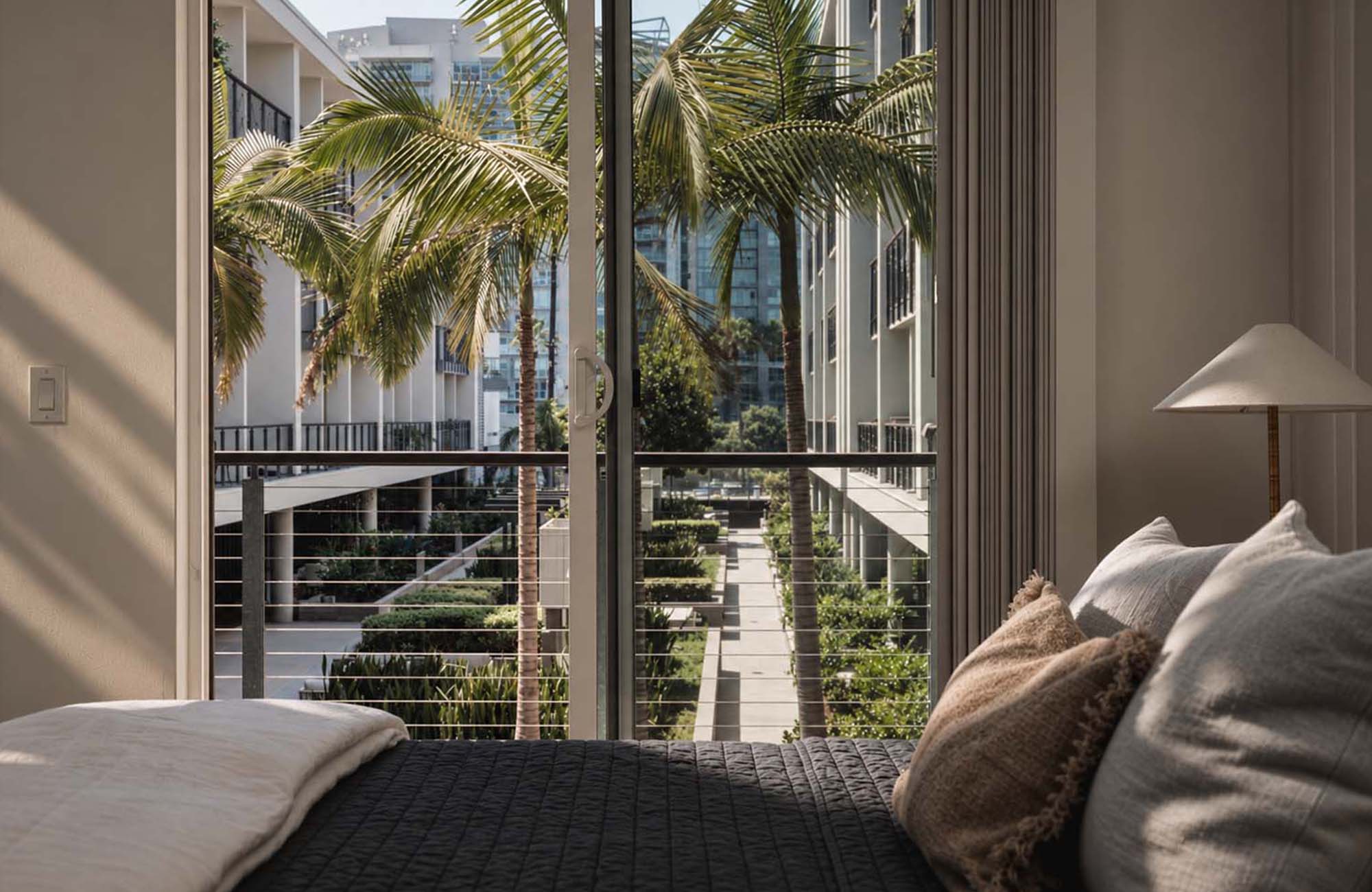 Apartment bedroom with sliding glass door opening to private balcony area with view of courtyard and palm trees.