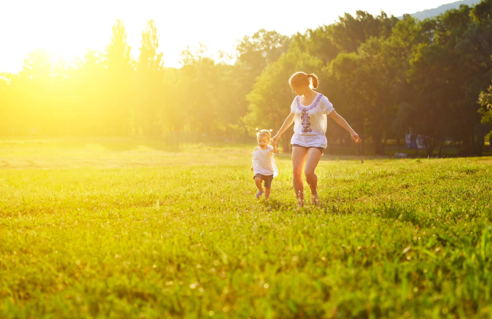 a mother in shorts & white blouse running in a field with her tiny daughter in Mill River Park - The Waypointe Apartments