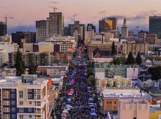 Aerial view of Oakland First Fridays at dusk, showing a vibrant crowd filling Telegraph Avenue with rows of colorful vendor tents, surrounded by mid-rise and high-rise buildings in downtown Oakland, with cranes and the Tribune Tower visible in the skyline.