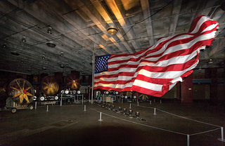 Several industrial fans keep a large American waving at an art exhibit