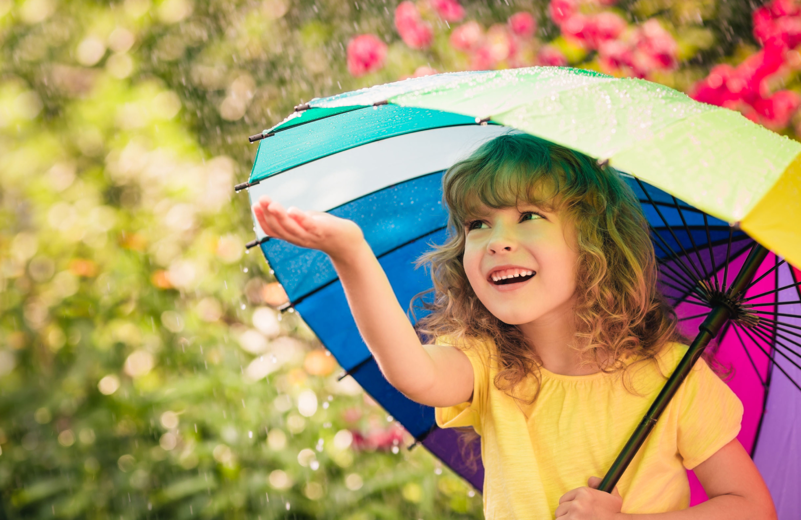 a little girl in a yellow shirt under a rainbow umbrella holding her hand out in the Rain in Norwalk