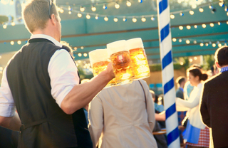 Guy in a black vest carrying several full beer mugs in one hand at an Octoberfest celebration.
