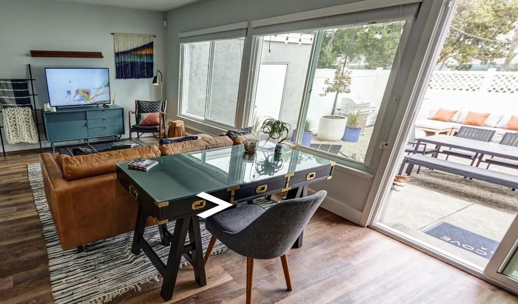 An apartment interior furnished with a desk, couch, coffee table, and media console, with a sliding door that opens to a large patio space.