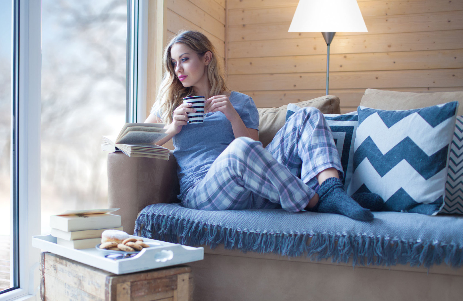 woman in pj pants relaxing on couch with a book & warm drink by a window - Hyde Square Apts