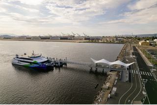 A San Francisco Bay Ferry docked at Alameda’s Seaplane Lagoon terminal on a calm day, with the waterfront, cranes, and bike-friendly pathways visible.