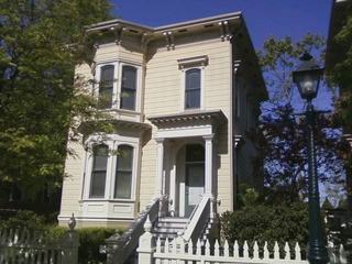  A restored two-story Victorian house at Preservation Park in Oakland, featuring cream-colored siding, bay windows, ornate trim, and a white picket fence framed by trees.