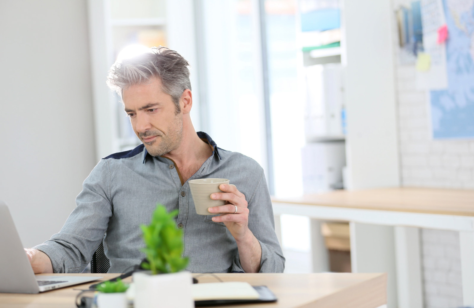a guy holding a coffee cup sitting at a desk in his home office working on laptop - Hyde Square apts