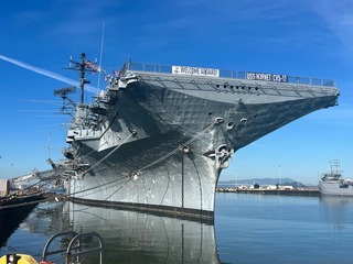 The USS Hornet aircraft carrier docked at Alameda Point on a clear day, with “Welcome Aboard! USS Hornet CVS-12” visible on the bow railing.