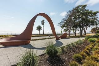 Sculptural wave-shaped bench in Shoreline Park, Alameda, California, positioned beside a walking path with palm trees and bay views in the background.