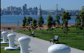 A waterfront path at Middle Harbor Shoreline Park in Oakland with benches, young trees, and white maritime bollards, looking out toward the San Francisco skyline and the Bay Bridge across the water.