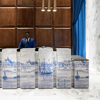 A smiling concierge stands behind the reception station in the 19 Dutch lobby. Marble floors, warm wood walls, and gold-tone lights.