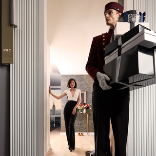 A bell boy in a burgundy suit carries a stack of boxes in the apartment's entryway with a sharply dressed woman in black and white jumpsuit behind him.