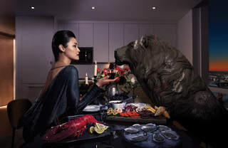 A woman dressed in black gazes into the eyes of a lion over a candle lit spread of lobster, charcuterie, and oysters.