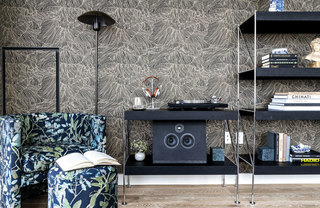 Apartment interior with a floral-patterned armchair and ottoman, a black floor lamp, and black media and book shelves, against a gray patterned wall.