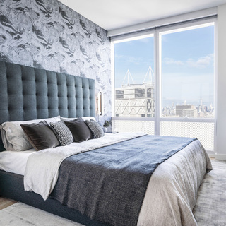 Bedroom interior. A king size bed with an oversized slate gray headboard and gray and white bedding, positioned in front of floor to ceiling windows with views of New York.