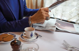 Close up of a man in blue suit sitting at a cafe table, reading a newspaper and drinking espresso.